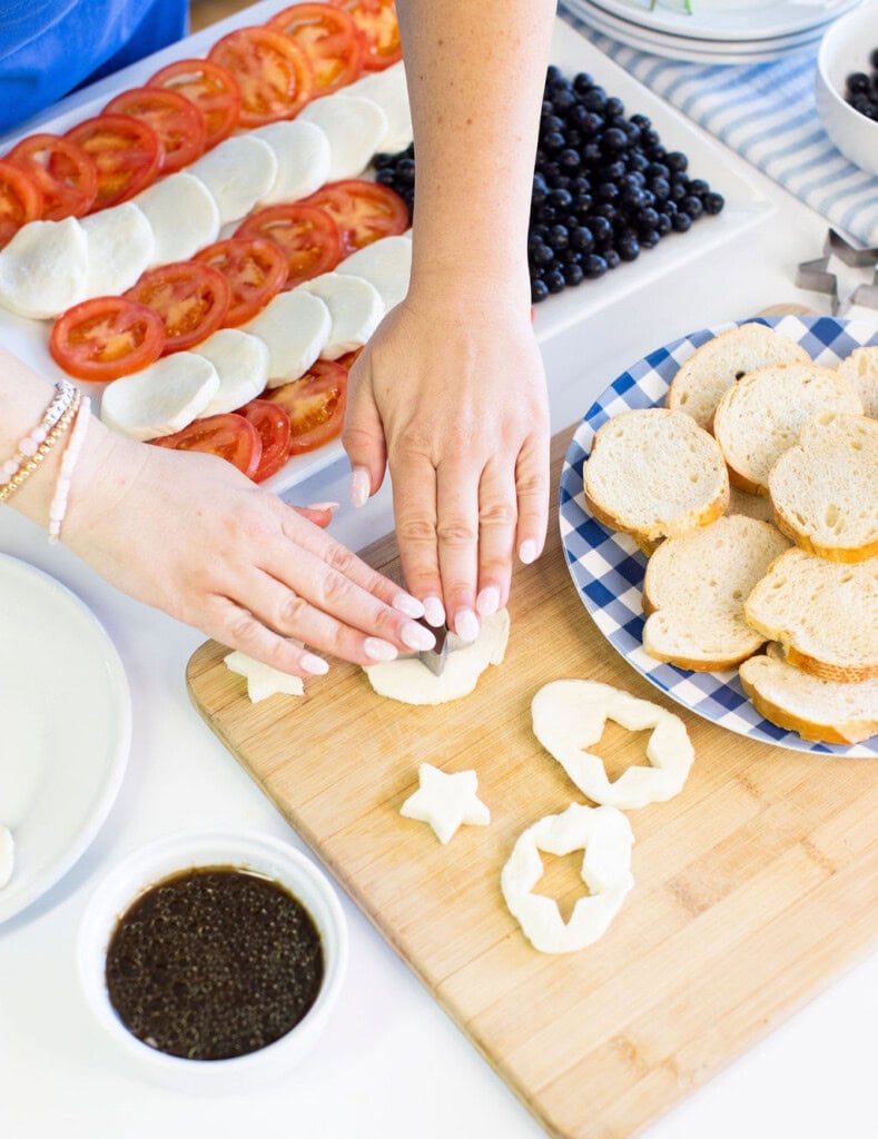 Two hands use cookie cutters to shape mozzarella cheese on a wooden board. Sliced bread, tomato-mozzarella stacks, blueberries, and a small bowl of dressing are visible on a bright kitchen table.