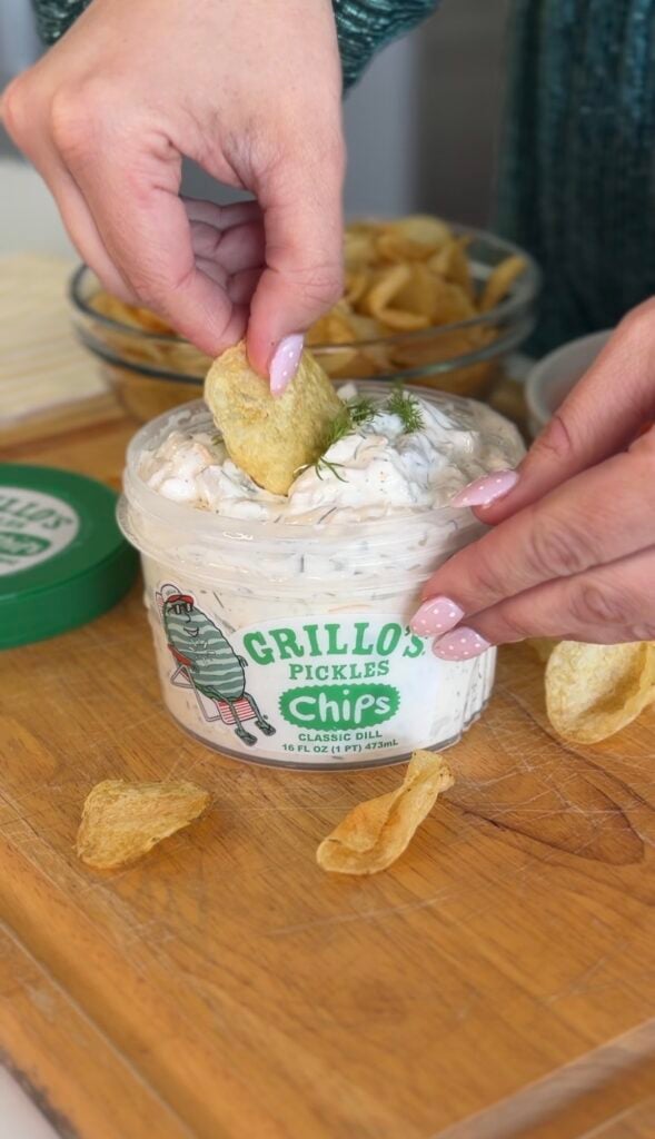 A person dips a potato chip into a tub of Grillos Pickles Chips dip on a wooden board, surrounded by more chips and snacks.