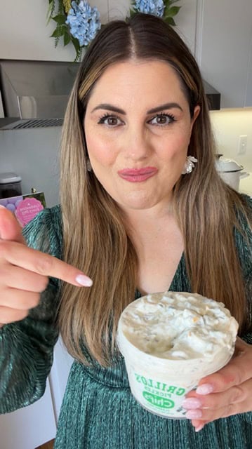A woman with long brown hair wearing a green dress stands in a kitchen, smiling and pointing at a tub of dip labeled “Grillo’s Pickle Chip Dip.” Blue flowers are visible in the background.