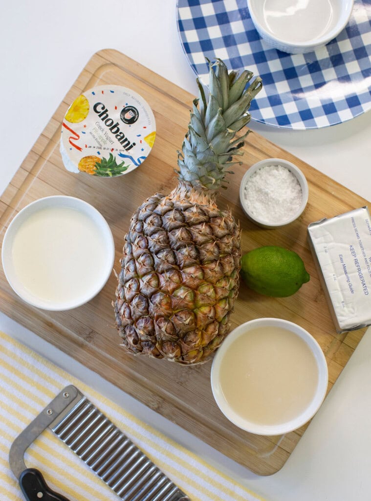 A whole pineapple, a lime, a block of cream cheese, a cup of yogurt, a bowl of cream, a bowl of sugar, and a bowl of powdered sugar on a wooden cutting board with a crinkle cutter and striped towel nearby.