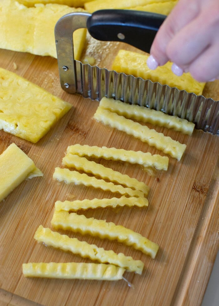 A hand using a crinkle cutter to slice pineapple into wavy strips on a wooden cutting board.