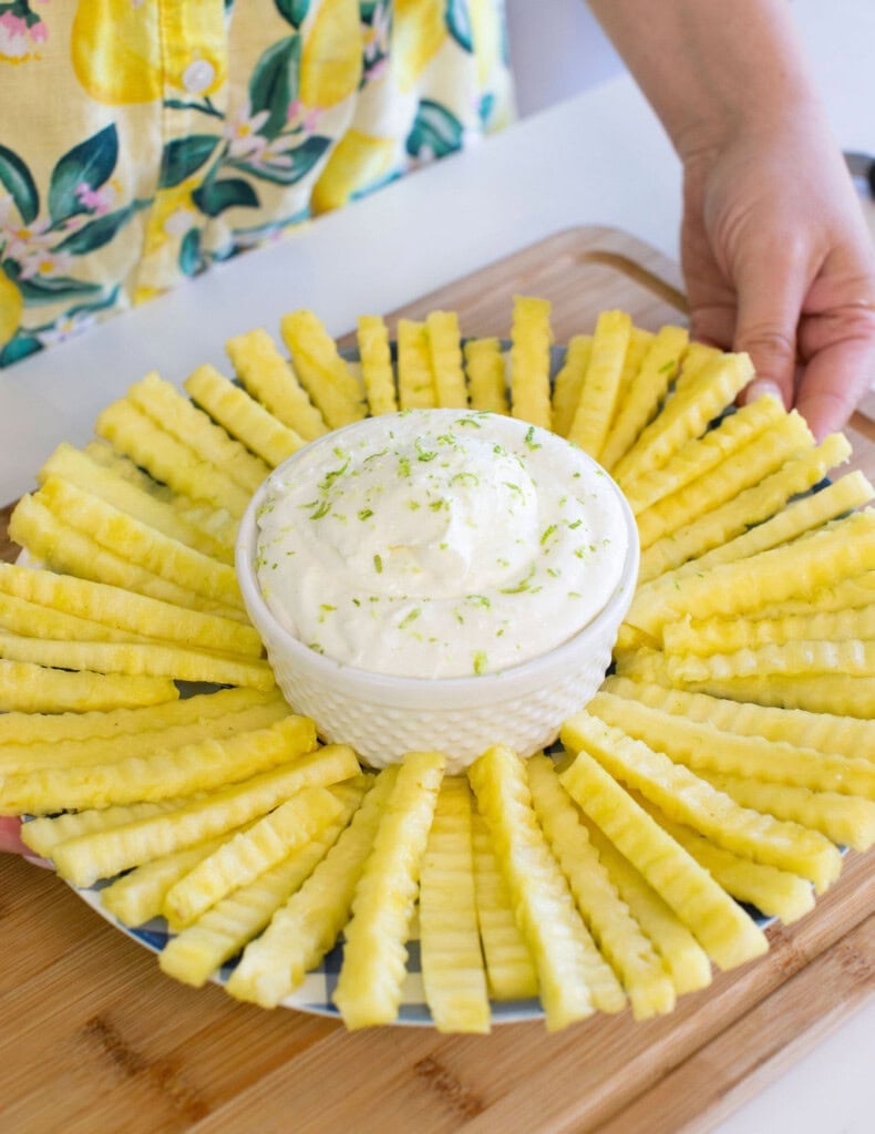 A person holds a platter of crinkle-cut pineapple slices arranged in a circle around a bowl of creamy dip, garnished with lime zest. The person is wearing a shirt with a yellow fruit and leaf pattern.