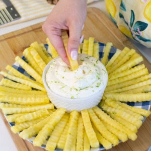 A hand dips a crinkle-cut pineapple stick into a bowl of creamy dip, surrounded by more pineapple sticks arranged in a circular pattern on a blue plate on a wooden board.