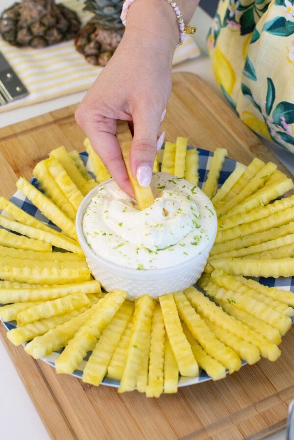 A hand dips a crinkle-cut pineapple stick into a bowl of creamy dip, surrounded by more pineapple sticks arranged in a circular pattern on a blue plate on a wooden board.