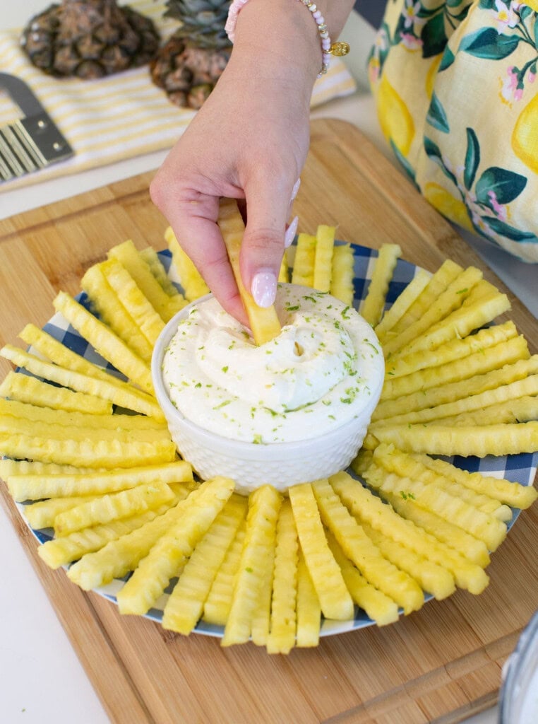 A hand dips a crinkle-cut pineapple stick into a bowl of creamy dip, surrounded by more pineapple sticks arranged in a circular pattern on a blue plate on a wooden board.