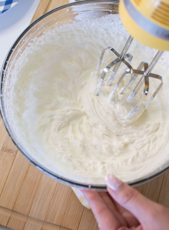 A hand holds a glass bowl of whipped cream being mixed with an electric hand mixer on a wooden surface.