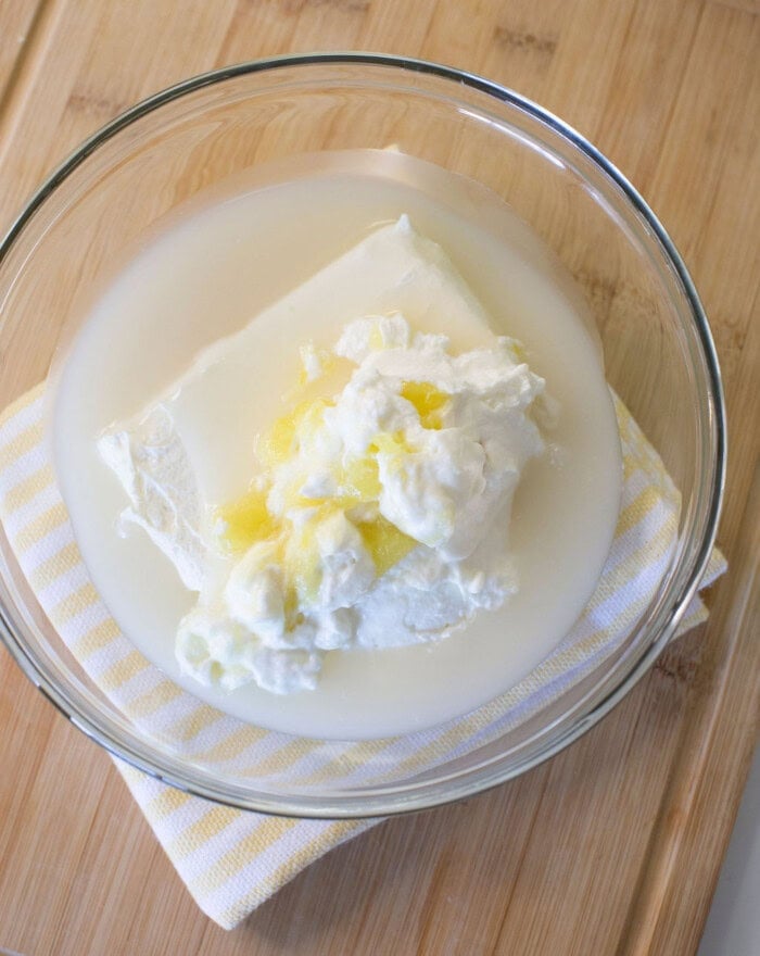 A glass bowl containing a block of cream cheese, lemon zest, and yogurt in liquid, placed on a striped cloth on a wooden surface.