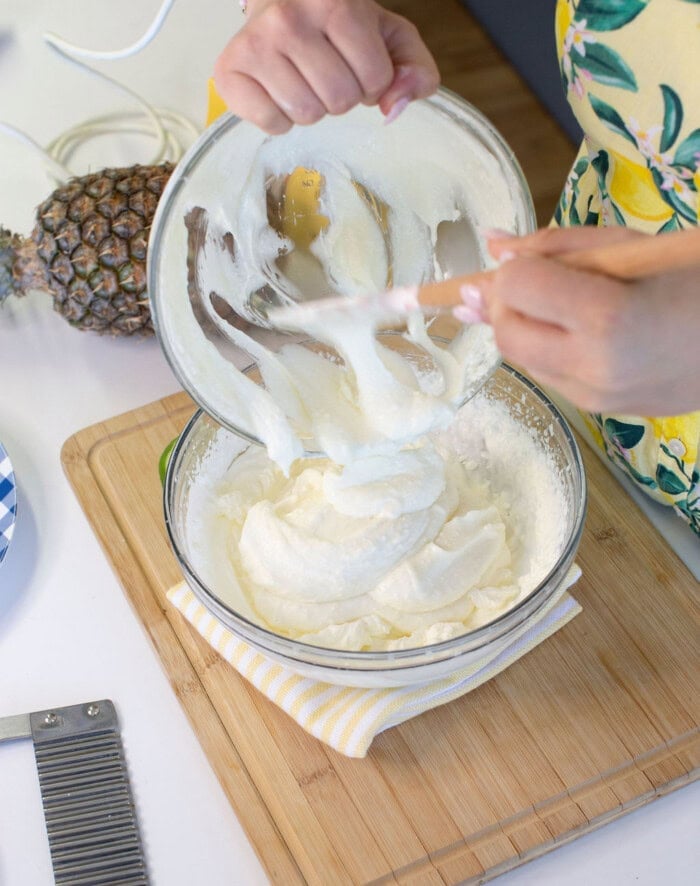 A person pours whipped cream from one glass bowl to another on a wooden cutting board. A pineapple, a striped cloth, and kitchen tools are nearby.