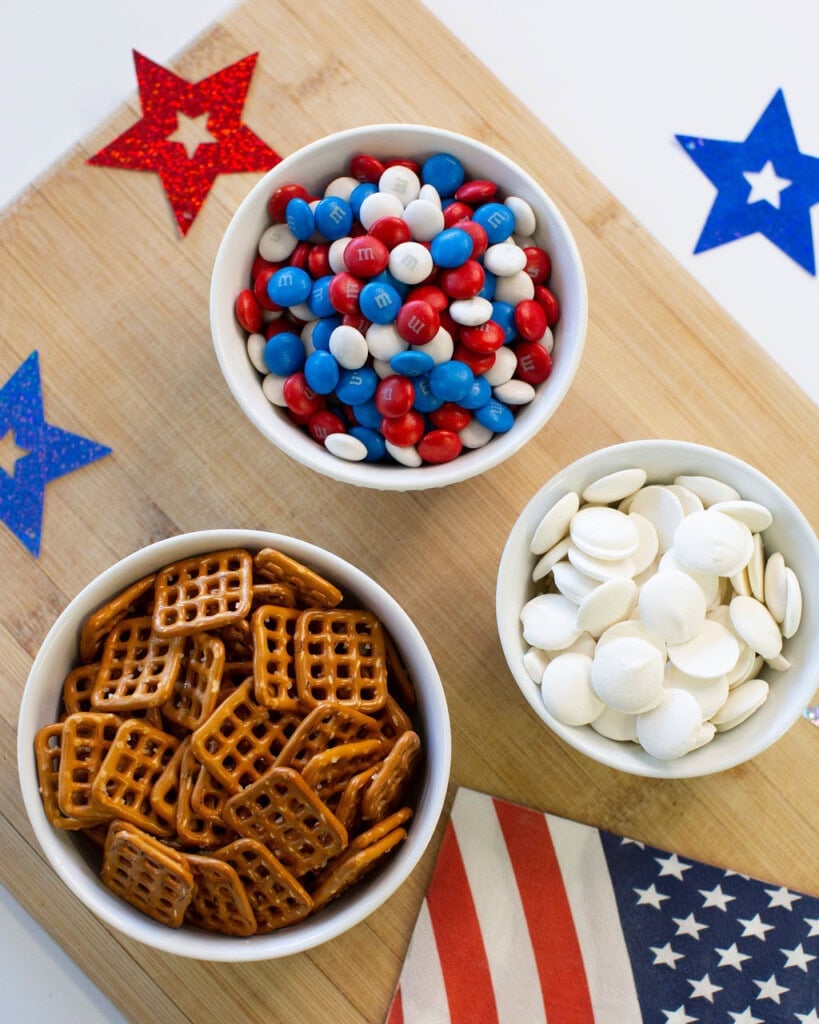 Three white bowls on a wooden board: one with red, white, and blue M&Ms, one with mini square pretzels, and one with white chocolate wafers. Patriotic star decorations and an American flag napkin are nearby.
