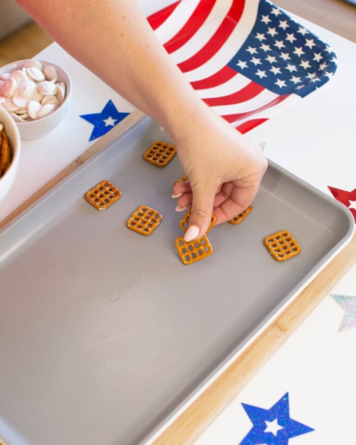 A hand places square pretzels on a baking sheet next to a bowl of candy melts, with an American flag napkin and colorful star decorations on the table.