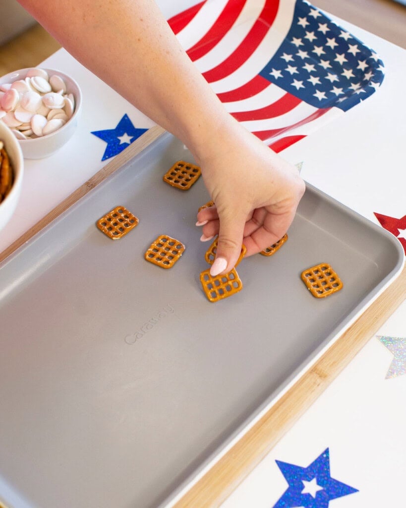 A hand places square pretzels on a baking sheet next to a bowl of candy melts, with an American flag napkin and colorful star decorations on the table.