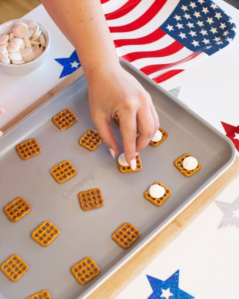 A hand places a white chocolate wafer on a square pretzel on a baking sheet. Other pretzels and wafers are arranged on the tray. An American flag and colorful star decorations are in the background.