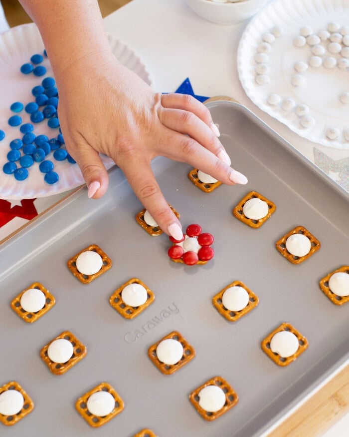 A hand arranges red candy pieces in a flower shape on pretzel squares topped with white chocolate, on a baking sheet with more pretzel treats and bowls of blue, red, and white candies nearby.