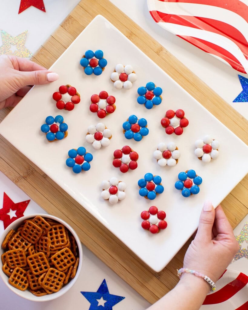 A person holds a white platter with flower-shaped treats made of red, white, and blue candies on square pretzels. A bowl of pretzels, star decorations, and an American flag plate are on the table.