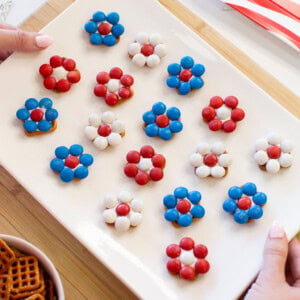 A white rectangular plate holds flower-shaped treats made of red, white, and blue candies on top of pretzels, arranged in rows; a bowl of pretzels and festive decor are nearby.