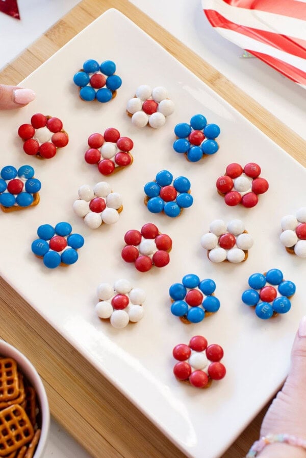 A white rectangular plate holds flower-shaped treats made of red, white, and blue candies on top of pretzels, arranged in rows; a bowl of pretzels and festive decor are nearby.
