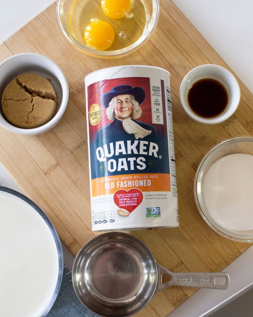 A container of Quaker Oats on a wooden board surrounded by eggs in a bowl, brown sugar, vanilla extract, granulated sugar, milk, and a measuring cup.