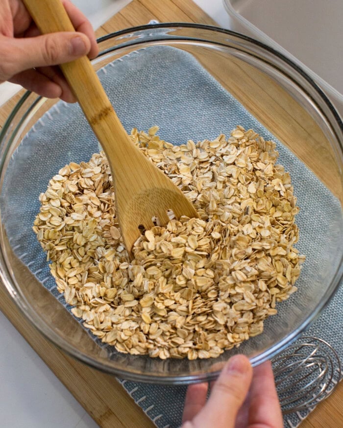 A person stirs rolled oats in a clear glass bowl with a wooden spoon, placed on a blue cloth and wooden board.