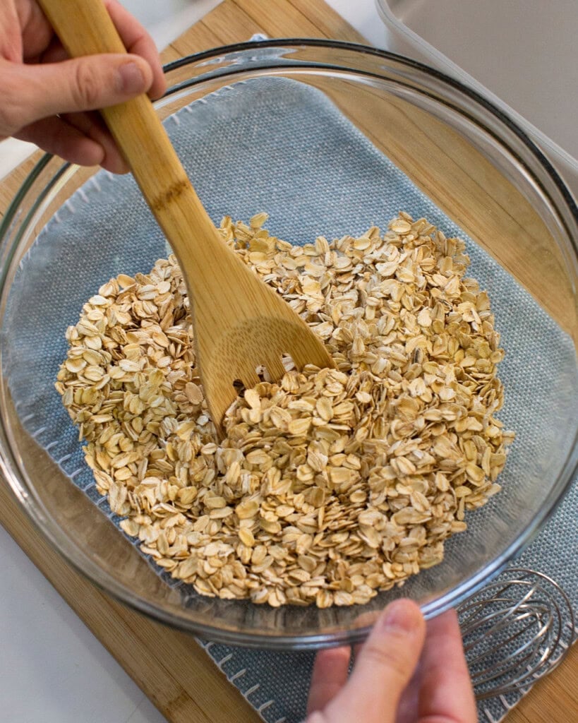 A person stirs rolled oats in a clear glass bowl with a wooden spoon, placed on a blue cloth and wooden board.