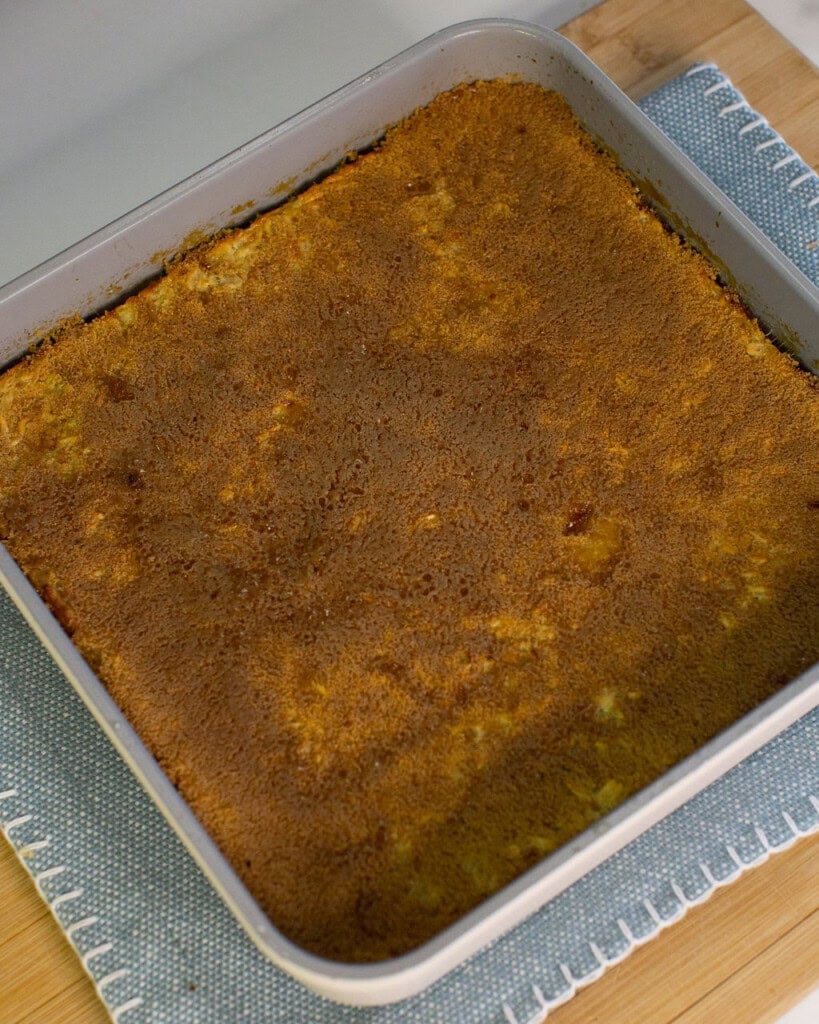 A square metal baking pan filled with a golden-brown baked dessert, possibly a cake or casserole, sits on a wooden surface with a blue and white placemat underneath.