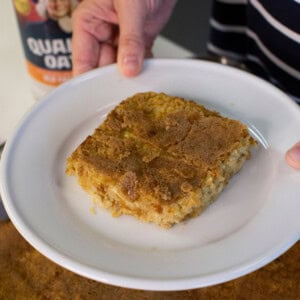 A person holds a white plate with a square slice of baked oatmeal dessert. A container of Quaker Oats is visible in the background on a counter.