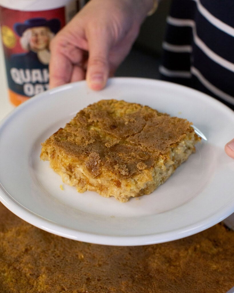 A person holds a white plate with a large square piece of baked oatmeal topped with a golden-brown crust. A container of Quaker Oats is visible in the background.