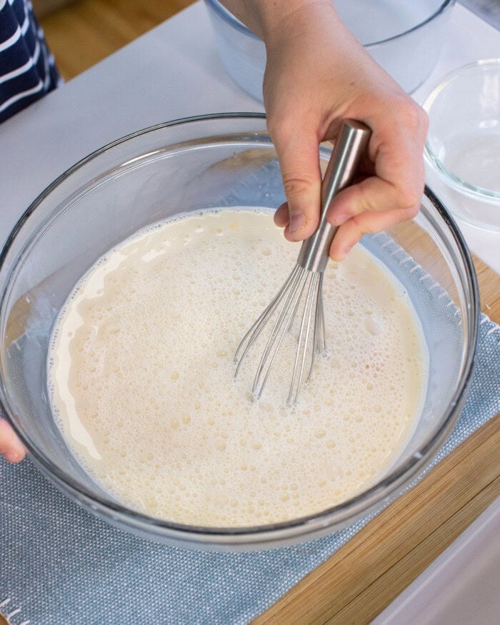 A person whisking a bowl of light, frothy batter or liquid mixture with a metal whisk on a table.