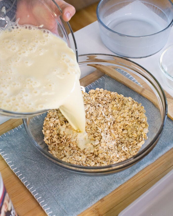 A close-up of a person pouring a creamy liquid from a glass measuring cup into a clear bowl filled with dry oats, with other bowls and kitchen items visible in the background.