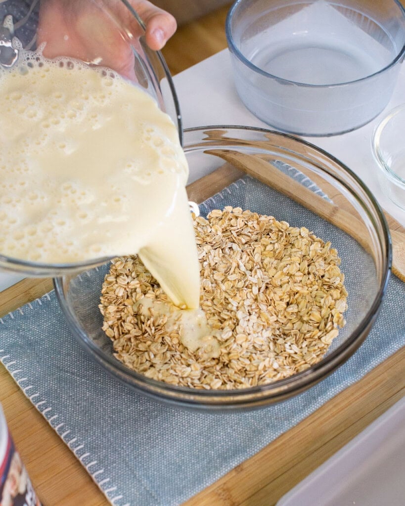 A close-up of a person pouring a creamy liquid from a glass measuring cup into a clear bowl filled with dry oats, with other bowls and kitchen items visible in the background.
