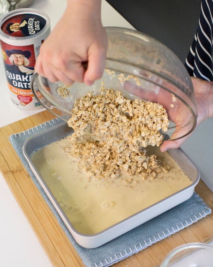 A person pours a bowl of uncooked oats into a square baking pan filled with liquid mixture. A Quaker Oats container sits on the counter nearby.