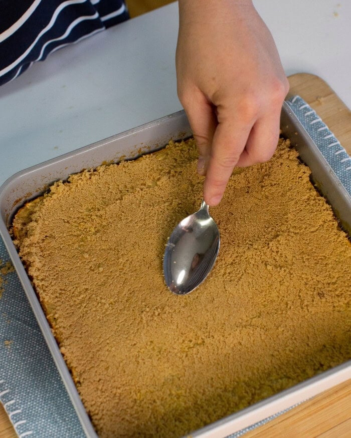 A hand holds a spoon, pressing down a layer of graham cracker crumbs in a square baking pan on a wooden surface. The edge of a blue and white cloth is visible underneath the pan.