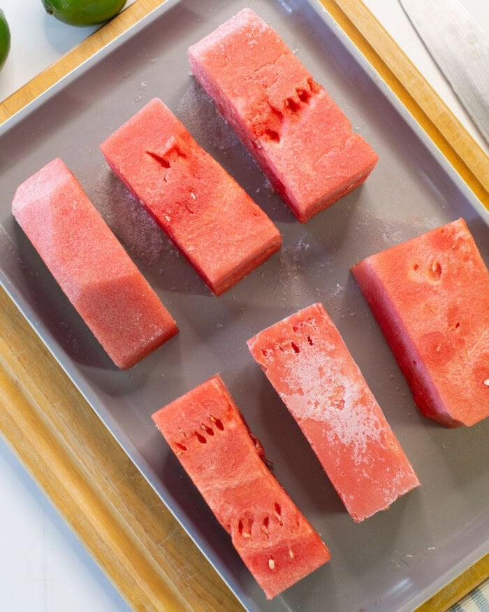 Six rectangular pieces of watermelon, some with visible frost, are arranged on a metal baking sheet. The tray sits on a wooden surface with part of a green lime visible in the background.