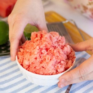 A person holds a white bowl filled with pink, shaved ice dessert on a blue and white striped cloth with a spoon and lime in the background.