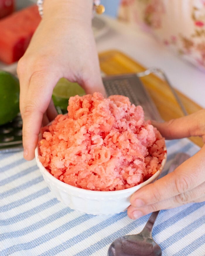 A person holds a white bowl filled with pink, shaved ice dessert on a blue and white striped cloth with a spoon and lime in the background.