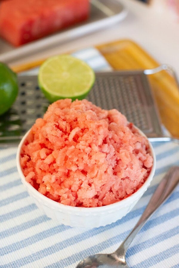 A bowl of pink watermelon granita sits on a striped cloth, with a spoon nearby. Behind it are a lime, a halved lime, a metal grater, and pieces of fresh watermelon on a tray.