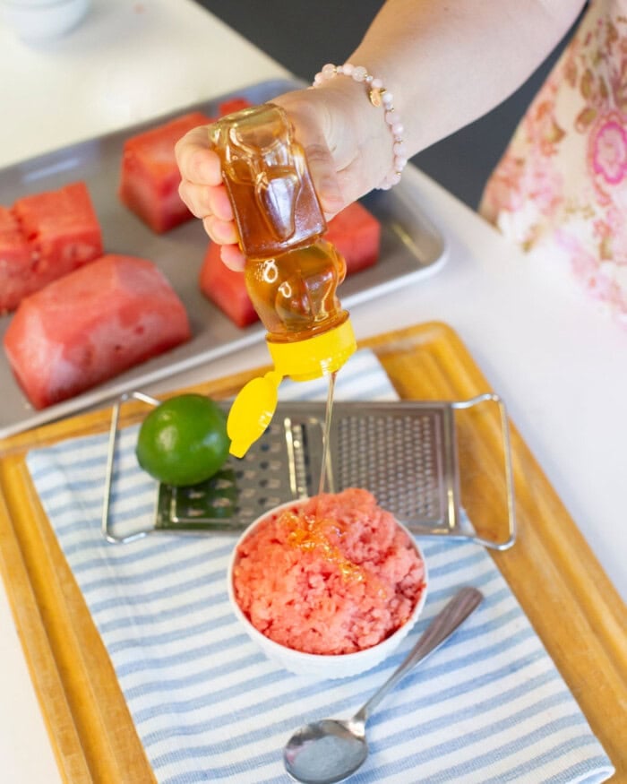 A hand drizzles honey from a squeeze bottle onto a bowl of watermelon granita, with a lime, metal grater, and watermelon chunks in the background on a tray. A spoon rests on a striped cloth nearby.
