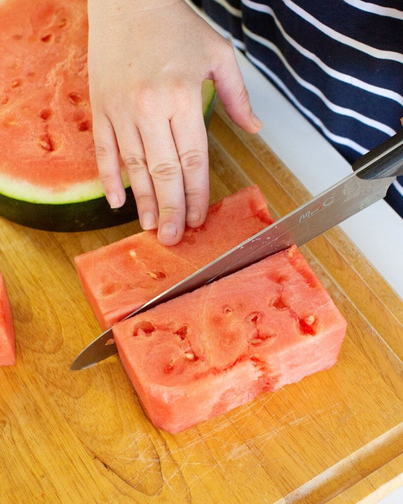 A hand holds a thick slice of watermelon on a wooden cutting board while a knife cuts through it; another half of watermelon is visible in the background.