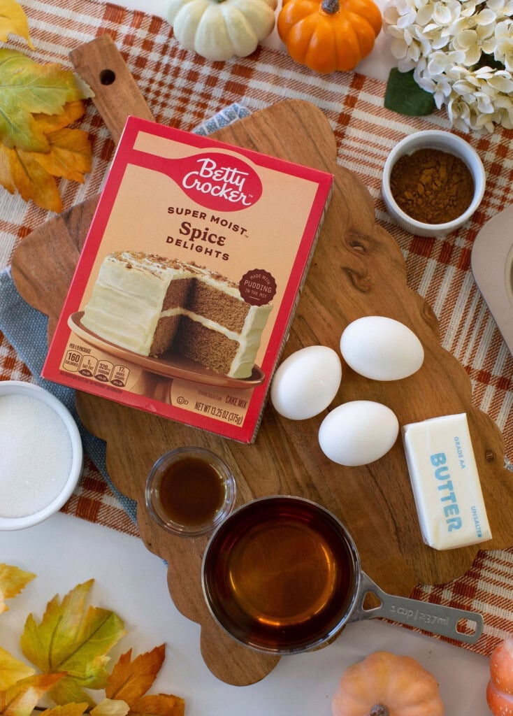 Cake mix box, eggs, butter, brown liquid in a measuring cup, sugar, and spices on a wooden board, surrounded by fall-themed decorations like pumpkins and leaves on a plaid tablecloth.