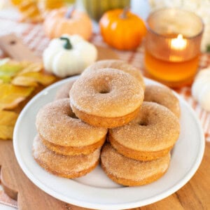 A plate of cinnamon sugar donuts sits on a wooden board. In the background, small pumpkins, fall leaves, and a lit candle create an autumn-themed scene.