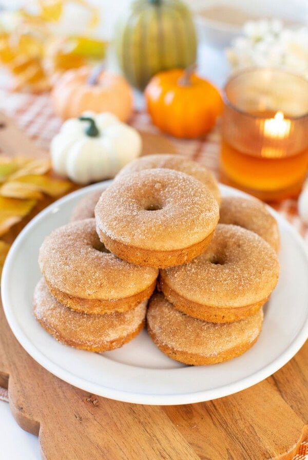A plate of cinnamon sugar donuts sits on a wooden board. In the background, small pumpkins, fall leaves, and a lit candle create an autumn-themed scene.