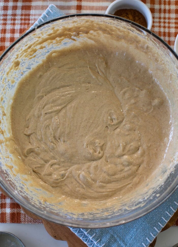 A glass bowl filled with light brown batter sits on a blue towel atop an orange plaid tablecloth, with small bowls nearby.