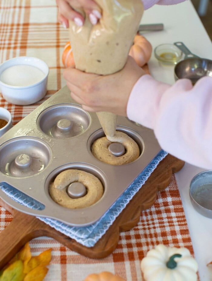 A person pipes batter into a donut pan on a wooden board, surrounded by baking ingredients and small pumpkins on a plaid tablecloth.