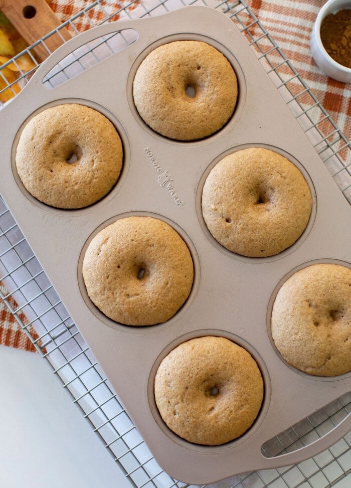 A six-cavity baking pan filled with baked donuts sits on a cooling rack over a plaid cloth. The donuts are light brown and evenly shaped.