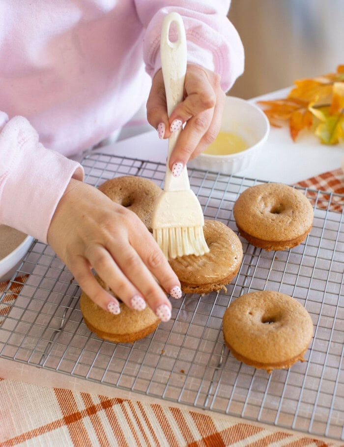 A person brushes glaze onto baked donuts on a cooling rack. Several donuts are visible, and the person has light pink, polka-dotted nails. A bowl and autumn leaves are in the background.