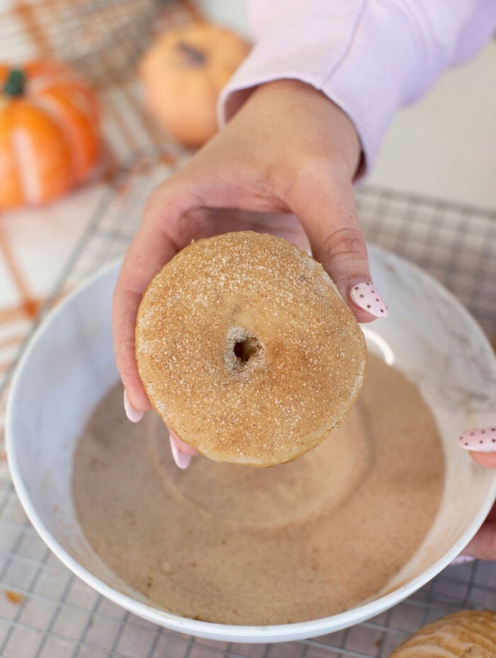 A hand holds a sugar-coated donut over a bowl of cinnamon sugar. A cooling rack and pumpkins are visible in the background.
