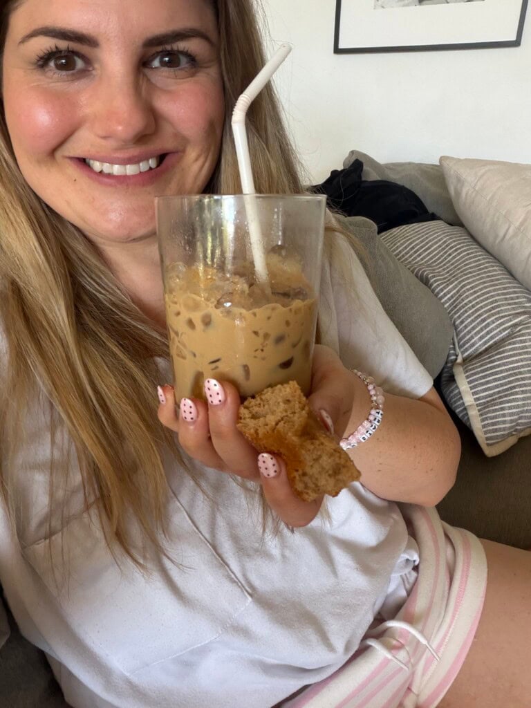 A woman smiles while holding an iced coffee with a white straw and a small piece of bread. She has light pink nails with white designs and is wearing a light top and striped shorts, sitting on a couch with grey pillows.