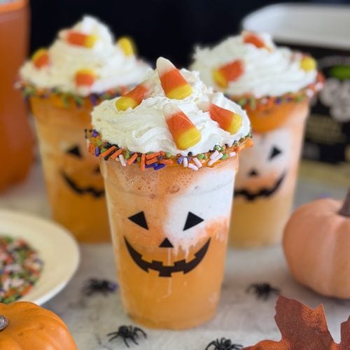 Three orange drinks in cups decorated with jack-o-lantern faces, topped with whipped cream, candy corn, and colorful sprinkles, surrounded by mini pumpkins, fake spiders, and autumn leaves.