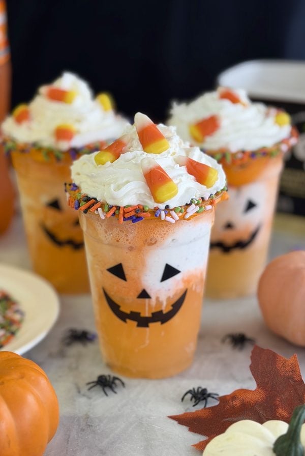 Three orange drinks in cups decorated with jack-o-lantern faces, topped with whipped cream, candy corn, and colorful sprinkles, surrounded by mini pumpkins, fake spiders, and autumn leaves.