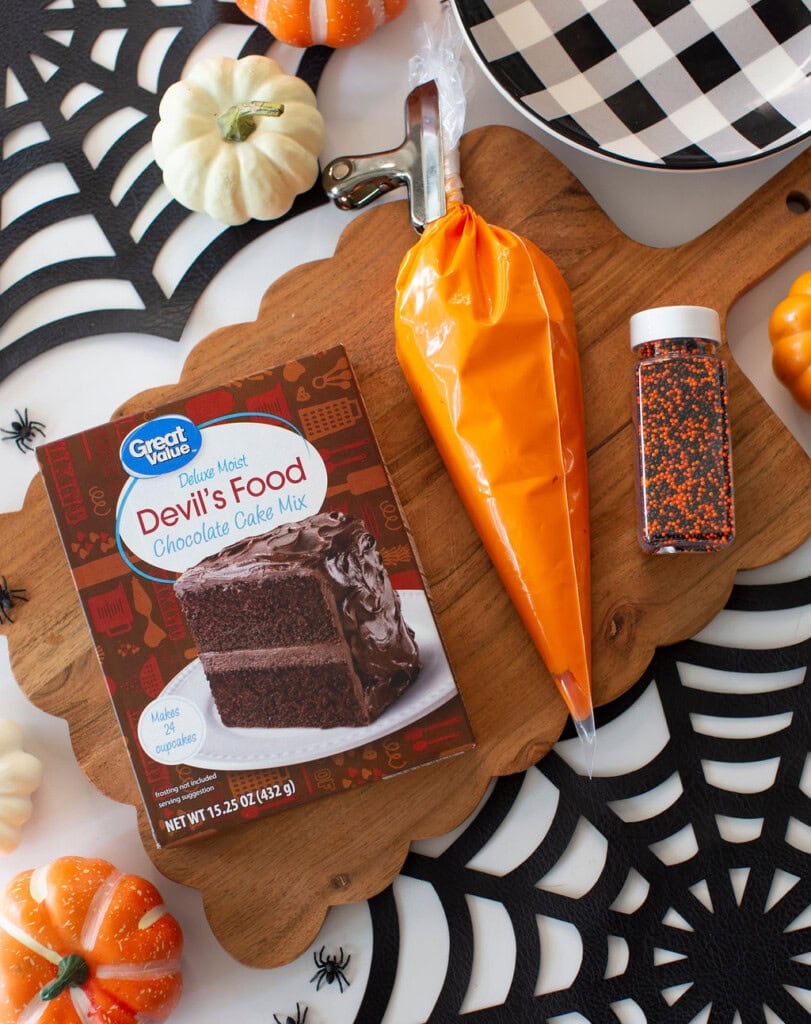 A box of Devils Food cake mix, orange icing in a piping bag, a bottle of black and orange sprinkles, and small pumpkins are arranged on a wooden board over a spiderweb placemat.