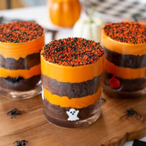Three layered Halloween desserts in glasses, topped with orange and black sprinkles. Each glass is decorated with small ghost and pumpkin figures. Mini plastic spiders are scattered on the wooden surface.
